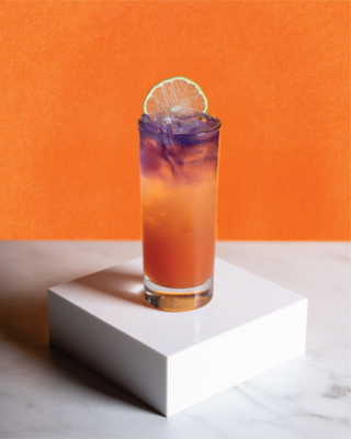A tall glass of iced purple and orange layered cocktail garnished with a lime wheel, displayed on a white pedestal against an orange background.