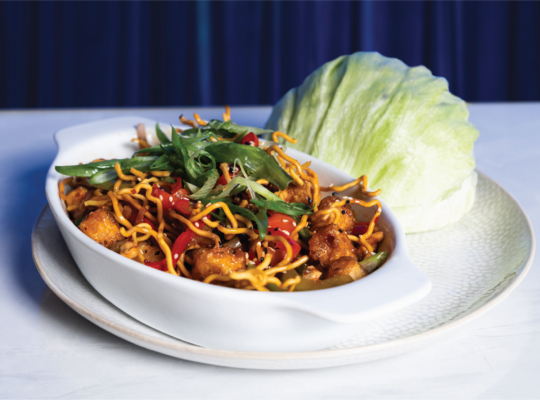 Spicy Asian chicken lettuce wraps garnished with crispy noodles and green onions, presented on a white plate against a blue curtain.