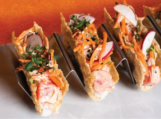 Three open-faced sushi tacos with salmon filling and vibrant vegetable toppings presented in a row against an orange background.